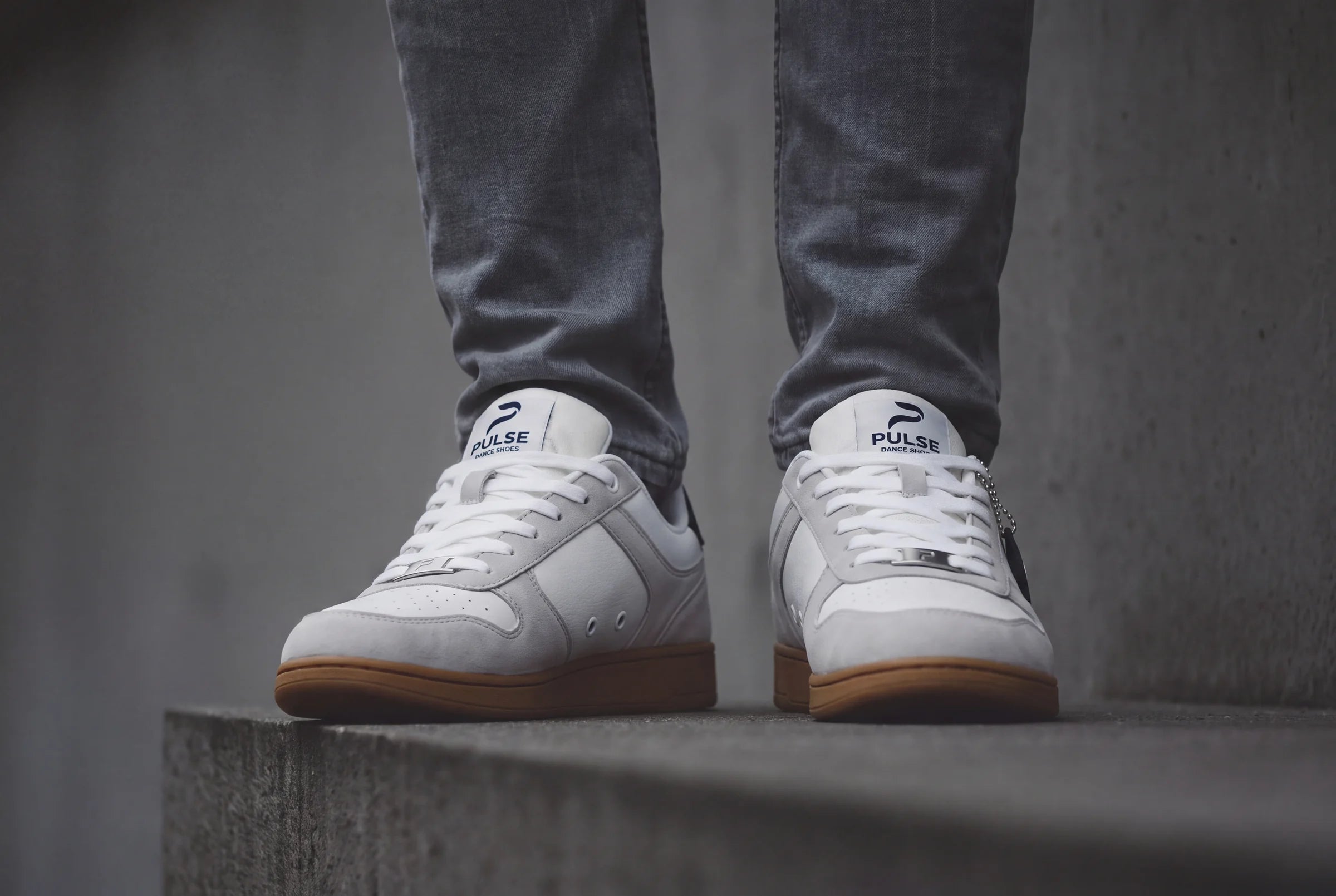 Feet in white dance sneakers with tan PU soles and gray jeans, standing on a concrete ledge
