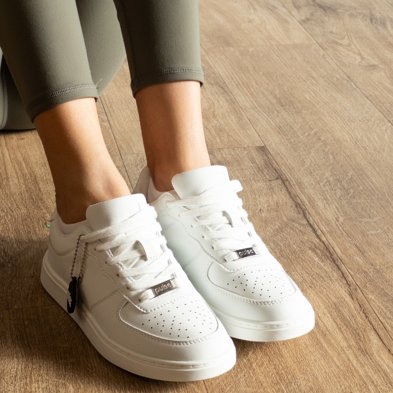 Woman's feet in white Pulse Dance Shoes dance sneakers with silver lace charms, olive leggings and a black hang tag