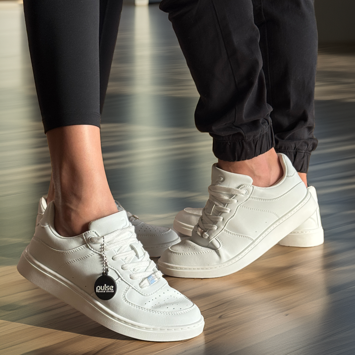Two people's feet in white low-top Pulse Dance Shoes dance sneakers on a polished wooden studio floor with a black hang tag